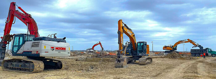 A construction site with four excavators digging and moving earth under a cloudy sky. A truck is parked on the right.